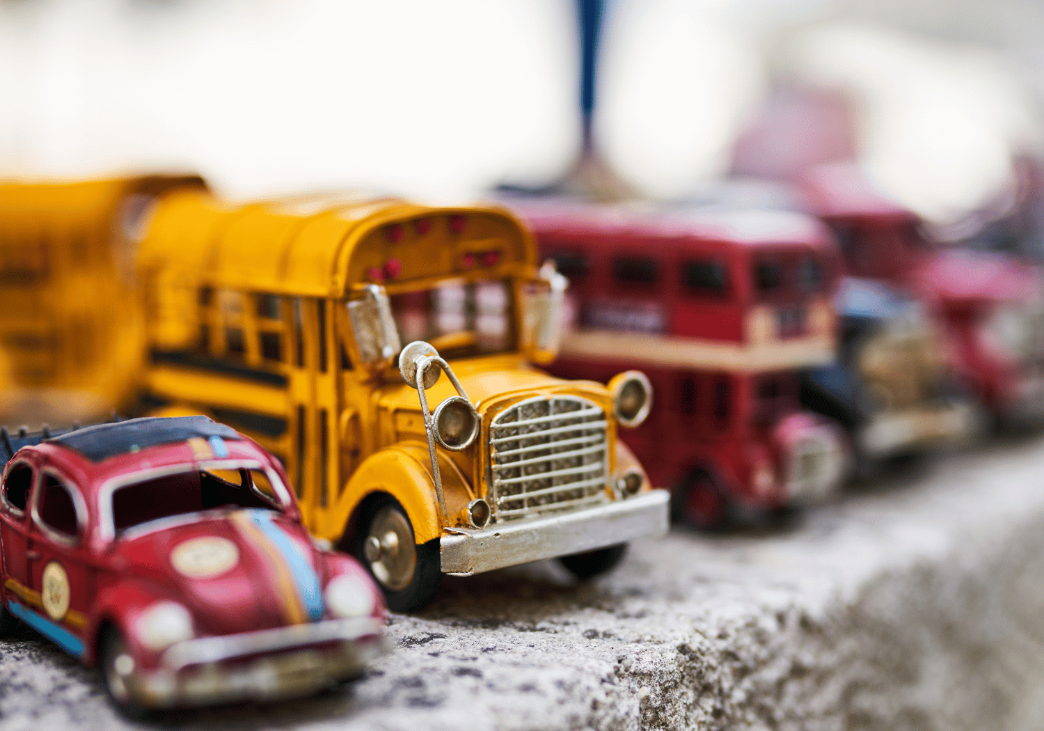 Close-up of a row of vintage toy cars on a rough surface, with a yellow school bus centered and a red car in the foreground; background vehicles are blurred.