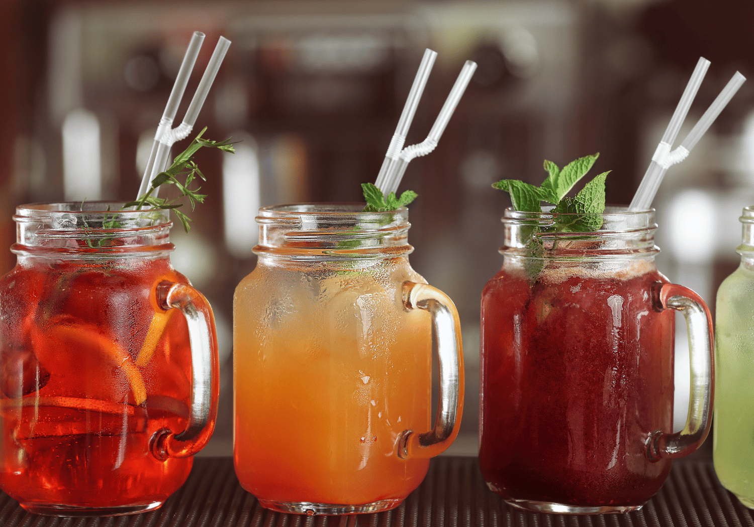 Three mason jar drinks with ice and mint, red/orange hues, on a bar counter with straws.