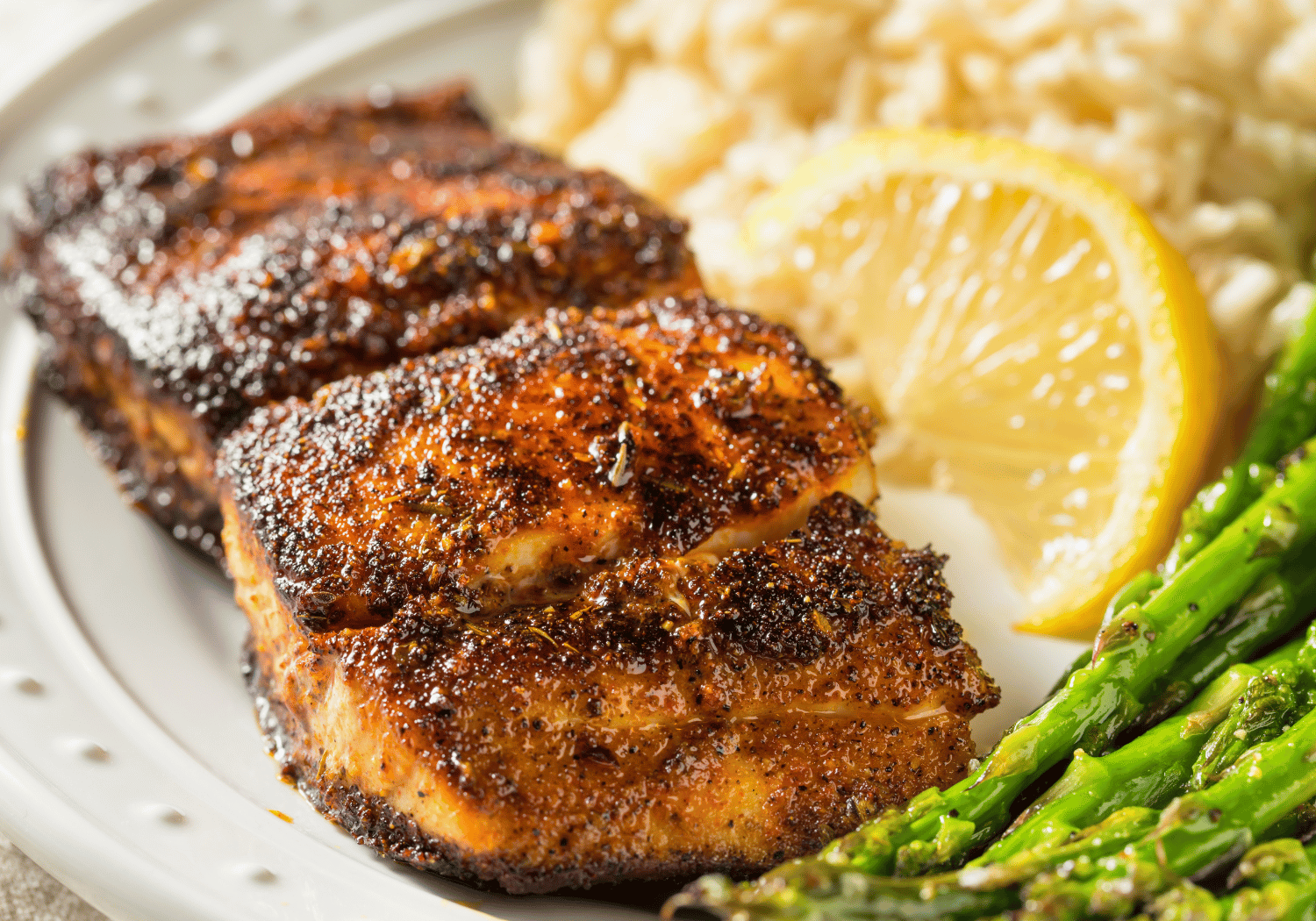 Two herb-crusted grilled chicken breasts on a plate with a lemon wedge, rice, and asparagus.